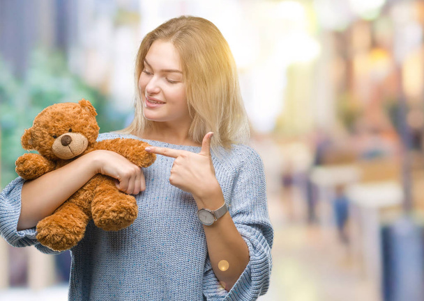 Молода біла жінка тримає милого плюшевого ведмедя над ізольованим тлом дуже щаслива, вказуючи рукою і пальцем
 - Фото, зображення
