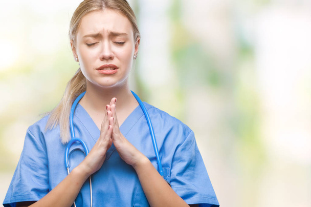 Young blonde surgeon doctor woman over isolated background begging and praying with hands together with hope expression on face very emotional and worried. Asking for forgiveness. Religion concept. - Φωτογραφία, εικόνα