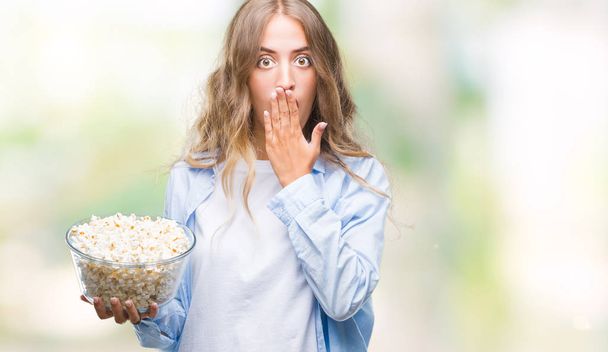 schöne junge blonde Frau, die Popcorn über isoliertem Hintergrunddeckel isst, Mund mit vor Scham geschockter Hand für Fehler, Ausdruck von Angst, Angst in der Stille, geheimes Konzept - Foto, Bild