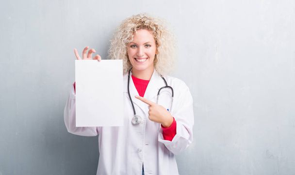Fiatal szőke doktor nő tartja üres papír lapot nagyon boldog, rámutatva a kéz és az ujjak grunge szürke falon - Fotó, kép