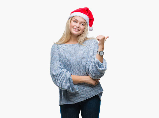 Mujer caucásica joven con sombrero de Navidad sobre fondo aislado sonriendo con la cara feliz mirando y señalando a un lado con el pulgar hacia arriba
. - Foto, Imagen
