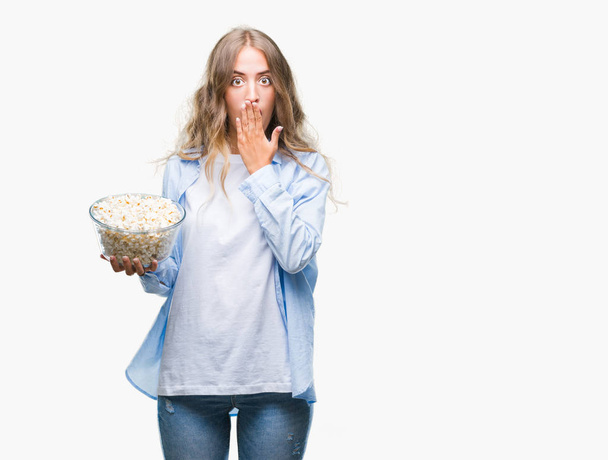 Bella giovane donna bionda che mangia popcorn su sfondo isolato coprire la bocca con la mano scioccata dalla vergogna per errore, espressione di paura, spaventata in silenzio, concetto segreto
 - Foto, immagini