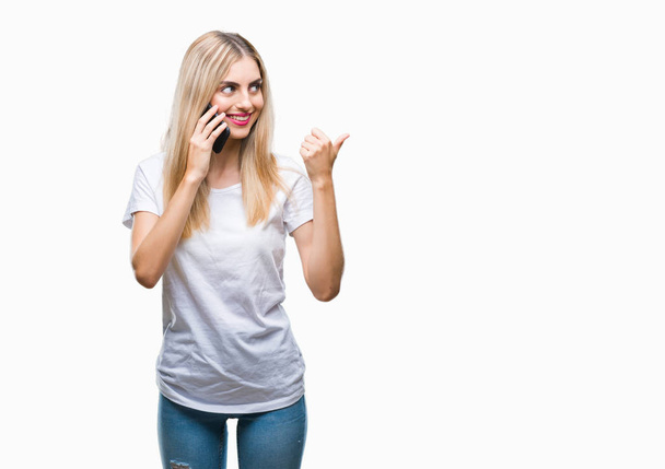 junge schöne blonde Frau spricht mit Smartphone über isolierten Hintergrund zeigt und zeigt mit dem Daumen zur Seite mit glücklichem Gesicht lächelnd - Foto, Bild