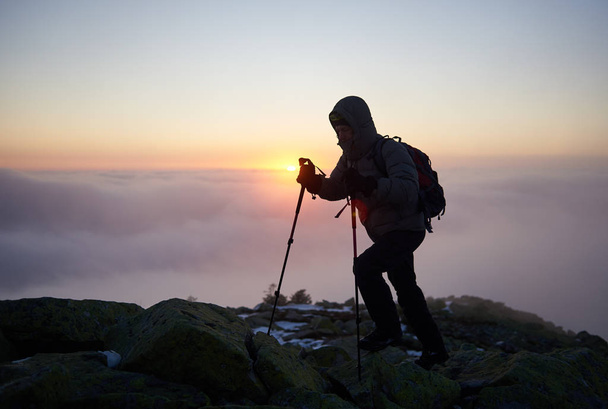 Silueta de excursionista turístico con mochila y bastones de trekking en pico rocoso de montaña con nubes de niebla en backgrond
 - Foto, Imagen