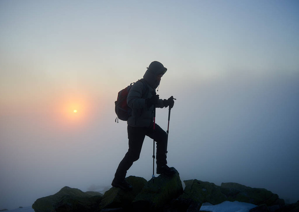 Silueta de excursionista turístico con mochila y bastones de trekking en pico rocoso de montaña con nubes de niebla en backgrond
 - Foto, Imagen