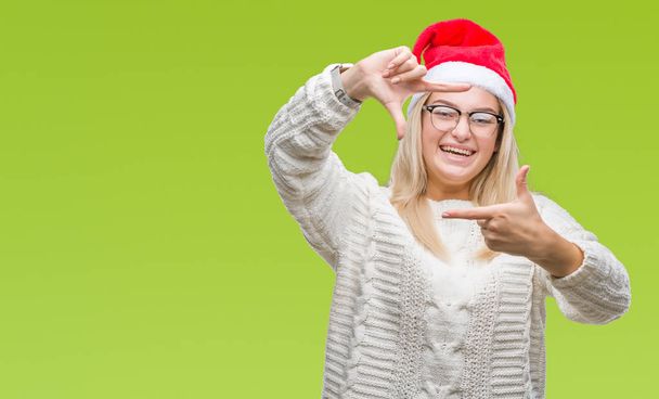 Mujer caucásica joven con sombrero de Navidad sobre fondo aislado sonriendo haciendo marco con las manos y los dedos con la cara feliz. Concepto de creatividad y fotografía
. - Foto, Imagen