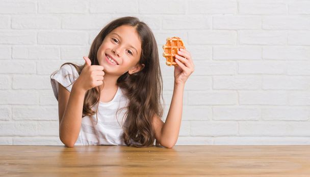 Young hispanic kid sitting on the table eating waffle happy with big smile doing ok sign, thumb up with fingers, excellent sign - Foto, Bild