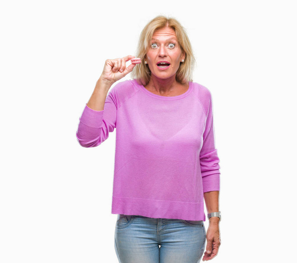 Mujer rubia de mediana edad comiendo galleta macaron rosa sobre fondo aislado asustado en shock con una cara sorpresa, asustado y excitado con expresión de miedo
 - Foto, Imagen