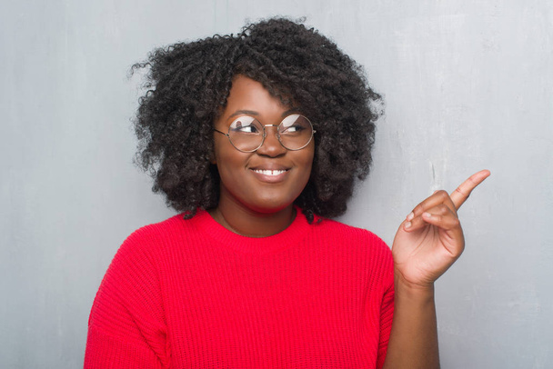 Jonge african american vrouw over grijs grunge muur dragen winter trui en glazen met een grote glimlach op het gezicht, met de hand en de vinger te wijzen naar de kant te kijken naar de camera. - Foto, afbeelding
