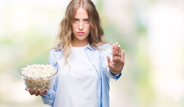 Kaunis nuori blondi nainen syö popcornia eristetyllä taustalla avoimella kädellä tekemällä stop-merkin vakavalla ja itsevarmalla ilmeellä, puolustus ele
 - Valokuva, kuva
