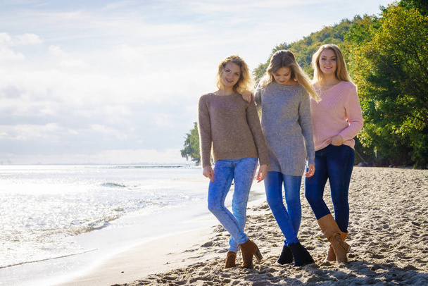 Three fashionable women wearing sweaters during warm autumnal weather spending their free time on sunny beach. Fashion models outdoor - Fotó, kép