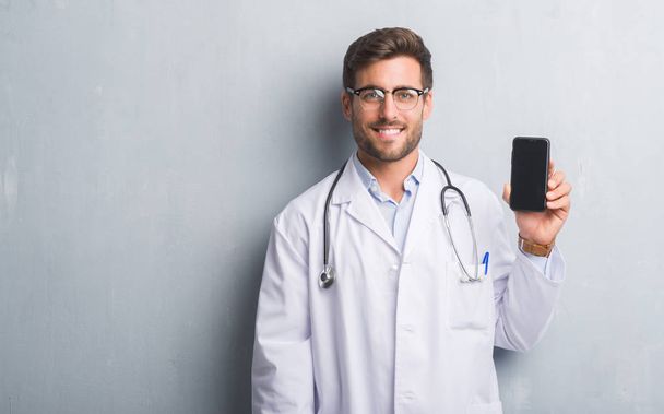 Beau jeune médecin homme sur le mur gris grunge tenant smartphone avec un visage heureux debout et souriant avec un sourire confiant montrant les dents
 - Photo, image