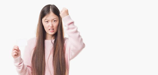 Młoda Chinka na białym tle gospodarstwa karty wizyty zirytowany i sfrustrowany krzycząc z gniewu, szalony i krzyczy z podniesiona ręka, gniew koncepcja - Zdjęcie, obraz