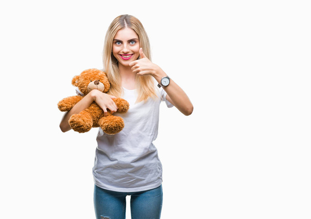 Young beautiful blonde woman holding teddy bear over isolated background happy with big smile doing ok sign, thumb up with fingers, excellent sign - Фото, изображение