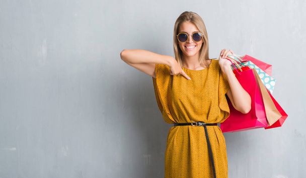 Hermosa mujer joven sobre la pared gris grunge sosteniendo bolsas de compras en las ventas con la cara sorpresa señalando el dedo a sí mismo
 - Foto, Imagen
