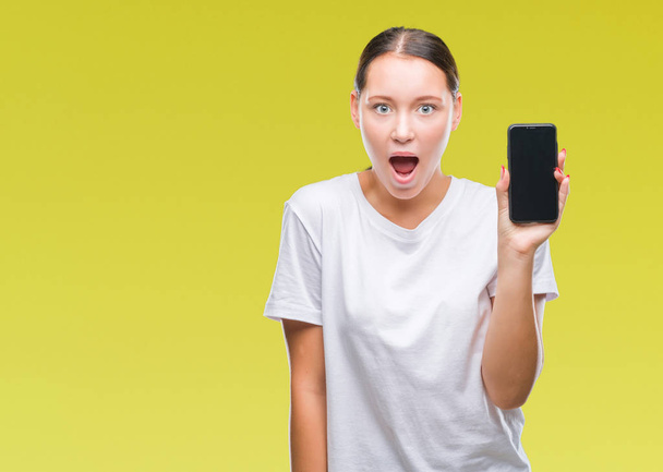 Young beautiful caucasian woman showing smartphone screen over isolated background scared in shock with a surprise face, afraid and excited with fear expression - Zdjęcie, obraz