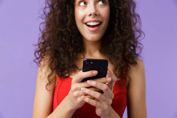 Imagem da jovem mulher 20s vestindo vestido vermelho segurando telefone celular preto de pé isolado sobre fundo violeta
 - Foto, Imagem