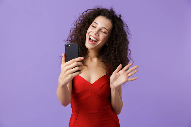 Image of joyous woman 20s wearing red dress using black smartphone standing isolated over violet background - Foto, Imagem