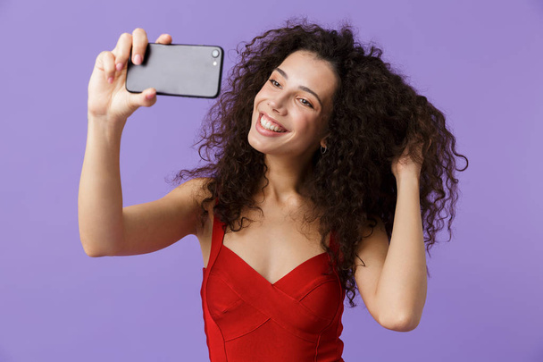 Image of curly woman 20s wearing red dress taking selfie photo on black smartphone standing isolated over violet background - Photo, Image