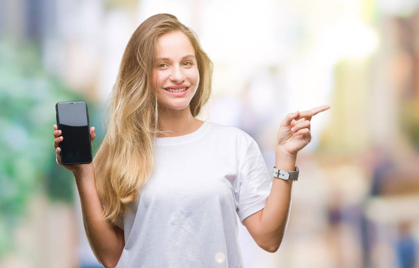 Jovem bela mulher loira mostrando tela do smartphone sobre fundo isolado muito feliz apontando com a mão e o dedo para o lado
 - Foto, Imagem