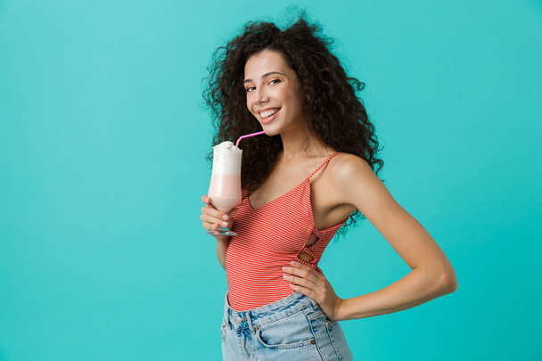 Photo of beautiful woman 20s with curly hair holding glass with cocktail isolated over blue background - Fotó, kép