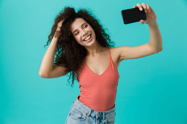 Photo of young woman 20s with curly hair taking selfie on mobile phone isolated over blue background - Foto, imagen