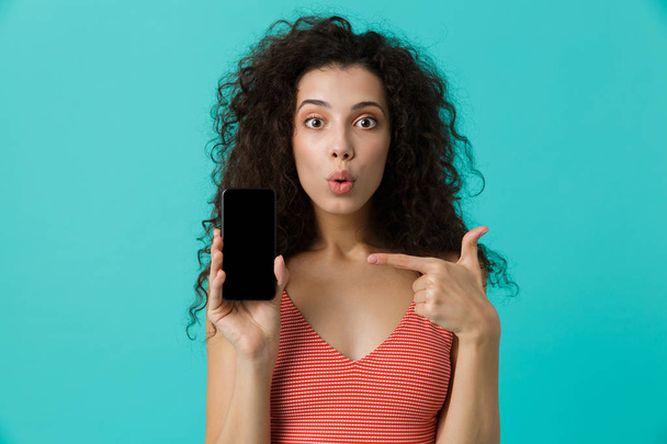 Photo of young woman 20s with curly hair pointing finger at mobile phone isolated over blue background - Fotografie, Obrázek