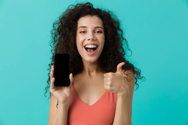 Photo of lovely woman 20s with curly hair smiling and holding cell phone isolated over blue background - Photo, image