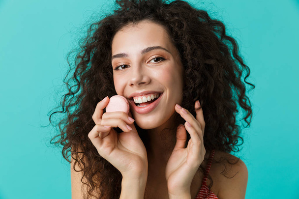 Photo closeup of joyful woman 20s with curly hair smiling and eating macaron biscuit isolated over blue background - Valokuva, kuva