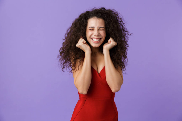 Portrait of a cheerful woman with dark curly hair wearing red dress isolated over violet background, celebrating success - Photo, Image