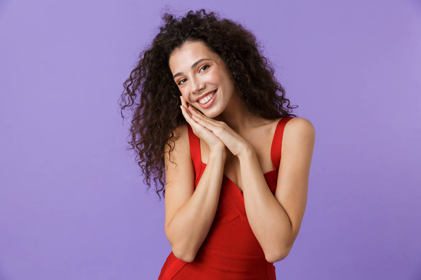 Portrait of a cheerful woman with dark curly hair wearing red dress isolated over violet background - Foto, immagini