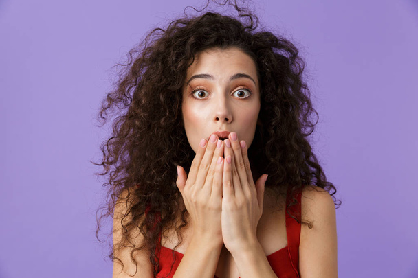Close up portrait of a shocked woman with dark curly hair wearing red dress isolated over violet background, covering face with hands - Φωτογραφία, εικόνα