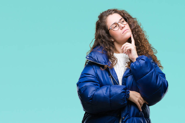 Beautiful young brunette curly hair girl wearing winter coat, glasses and sweater over isolated background with hand on chin thinking about question, pensive expression. Smiling with thoughtful face. Doubt concept. - Фото, зображення