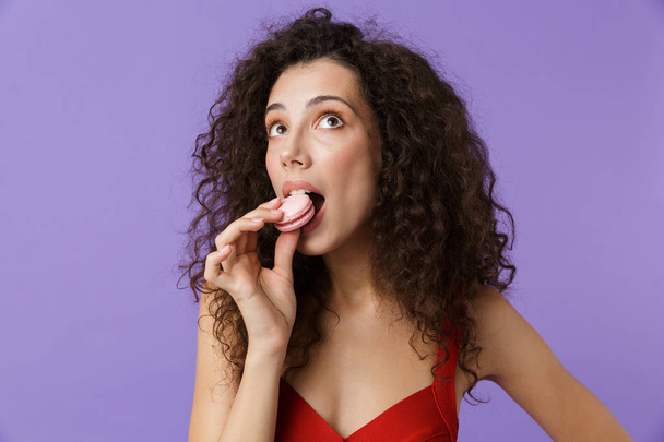 Portrait of a lovely woman with dark curly hair wearing red dress isolated over violet background, eating macaroon - Foto, Imagen