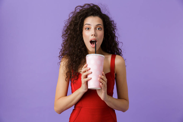 Portrait of a cheerful woman with dark curly hair wearing red dress isolated over violet background, drinking from a plastic cup with a straw - Foto, Imagem