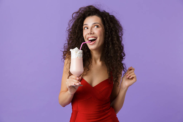 Portrait of an excited woman with dark curly hair wearing red dress isolated over violet background, drinking milk shake forom a glass - Photo, Image
