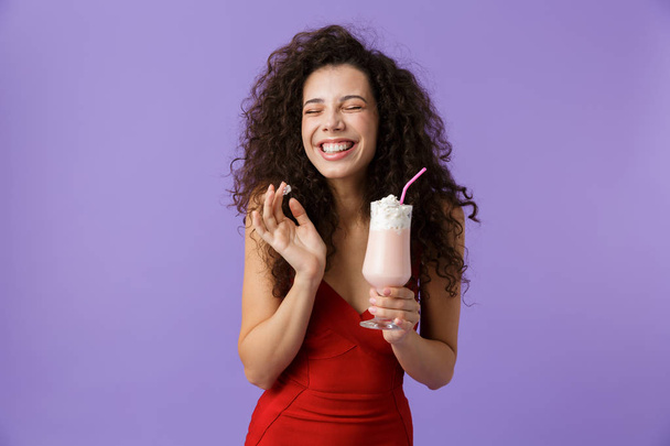 Retrato de uma mulher satisfeita com cabelos encaracolados escuros usando vestido vermelho isolado sobre fundo violeta, bebendo batido de leite de um copo
 - Foto, Imagem