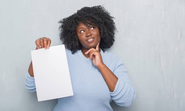 Young african american woman over grey grunge wall holding blank paper sheet serious face thinking about question, very confused idea - Fotoğraf, Görsel