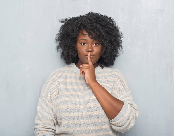 Nuori afrikkalainen amerikkalainen plus kokoa nainen harmaan grunge seinä yllään villapaita pyytää olla hiljaa sormi huulilla. Hiljaisuus ja salainen käsite
. - Valokuva, kuva