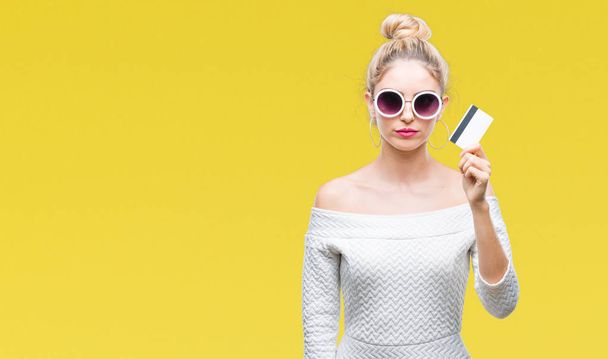Young beautiful blonde woman holding credit card over isolated background with a confident expression on smart face thinking serious - Foto, immagini