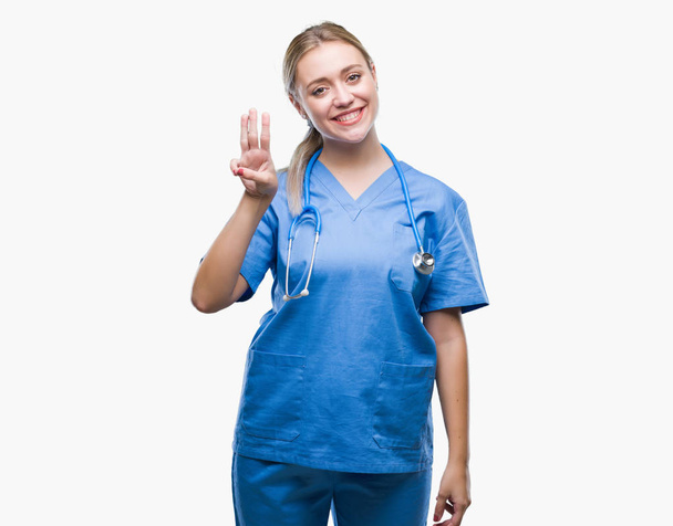 Genç Sarışın cerrah doktor kadın gösterilen ve parmakları ile işaret izole arka plan yüzünden kendinden emin ve mutlu gülümseyerek iken üçüncü. - Fotoğraf, Görsel