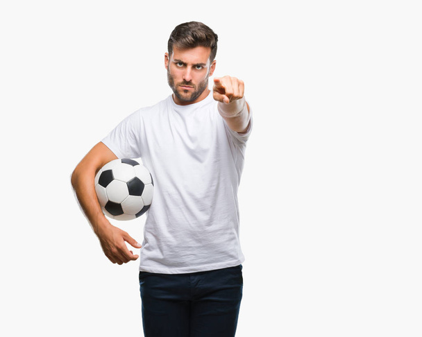 Όμορφος νεαρός, κρατώντας την μπάλα ποδοσφαίρου ποδοσφαίρου πάνω από απομονωμένη φόντο δείχνοντας με το δάχτυλο στη φωτογραφική μηχανή και σε εσάς, το χέρι συνδεθείτε, θετική και σίγουρη κίνηση από το μέτωπο - Φωτογραφία, εικόνα