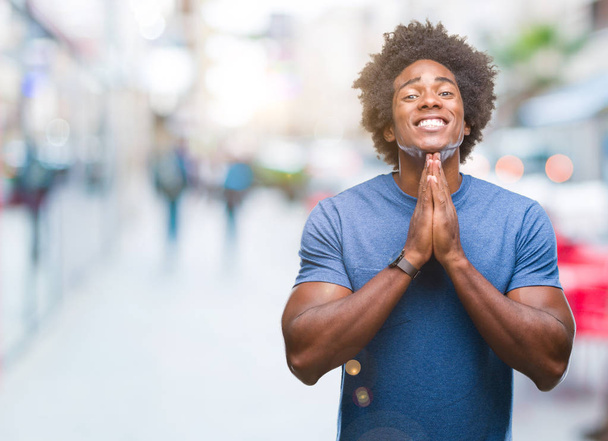 Hombre afroamericano sobre fondo aislado rezando con las manos juntas pidiendo perdón sonriendo confiado
. - Foto, imagen