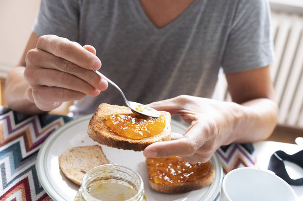 крупним планом молодий кавказький чоловік, одягнений у казуальну сіру футболку, сидить за столом, розкладаючи апельсинове або персикове варення на тості
 - Фото, зображення