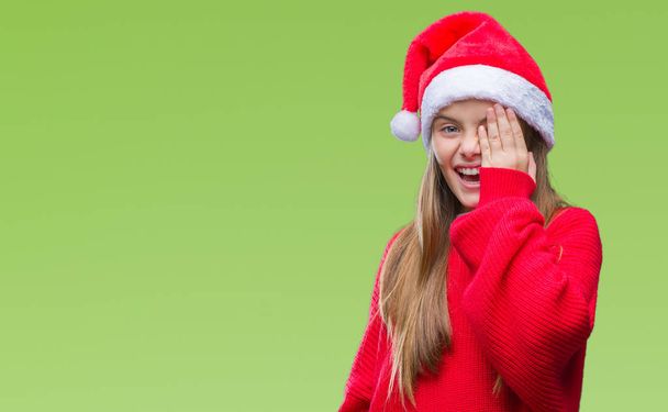 Nuori kaunis tyttö yllään joulu hattu yli eristetty tausta peittää toisen silmän käsin luottavainen hymy kasvoilla ja yllätys tunteita
. - Valokuva, kuva
