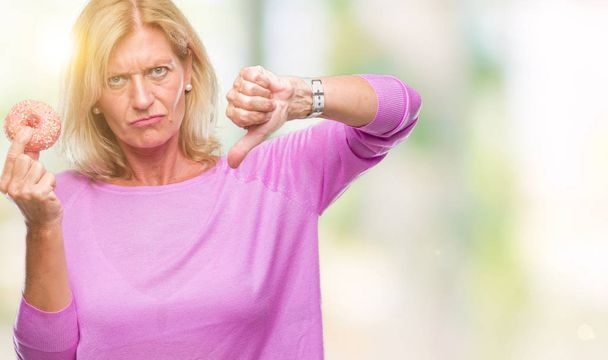 Femme blonde du moyen âge mangeant un beignet rose sur fond isolé avec un visage en colère, signe négatif déplaisant avec les pouces baissés, concept de rejet
 - Photo, image