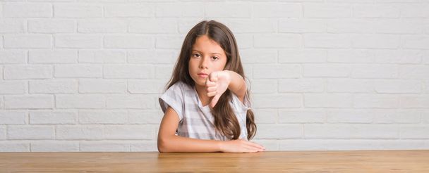 Fiatal spanyol gyerek ül az asztal otthon látszó boldogtalan és dühös mutatja elutasítása, és negatív, a hüvelykujj le gesztus. Rossz kifejezés. - Fotó, kép