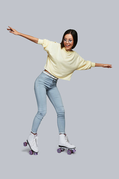Full length of young Asian woman smiling while skating against grey background - Photo, image