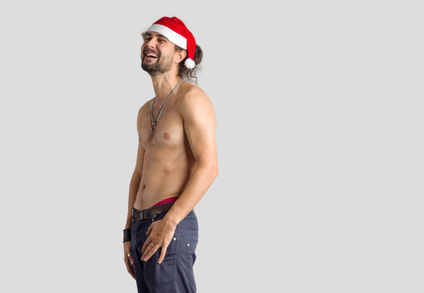 junger Mann mit nacktem Oberkörper und roter Weihnachtsmütze auf grauem Hintergrund. - Foto, Bild