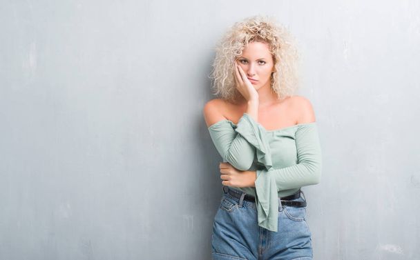 Young blonde woman with curly hair over grunge grey background thinking looking tired and bored with depression problems with crossed arms. - Photo, Image